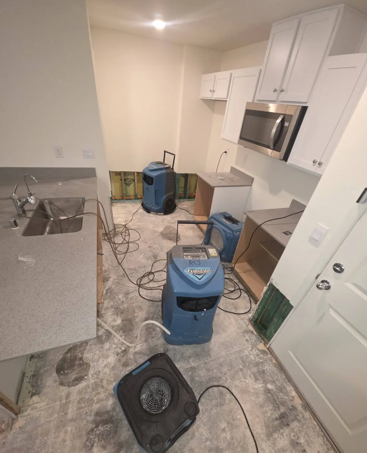 A kitchen with a dehumidifier and air movers set up on a bare floor