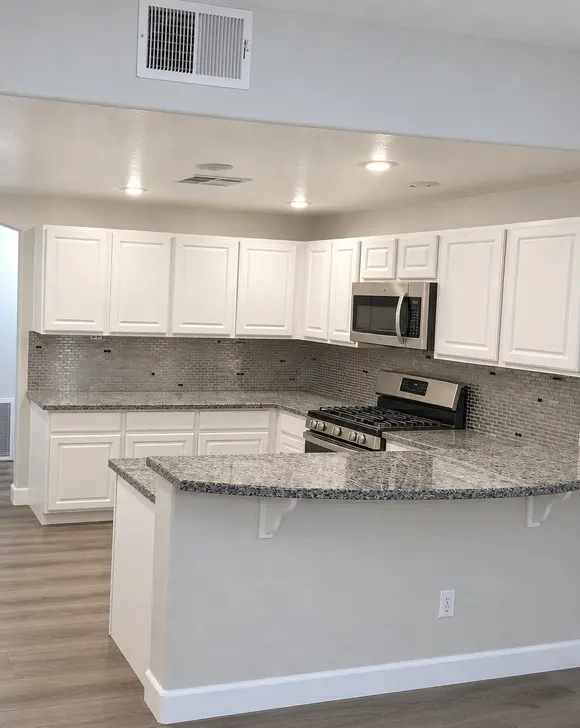 A white kitchen with a curved grey granite peninsula, a silver mosaic backsplash and stainless appliances